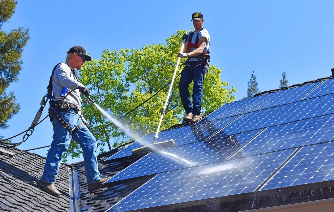 Two solar panel cleaning technicians cleaning solar panels with a brush and hose on a rooftop